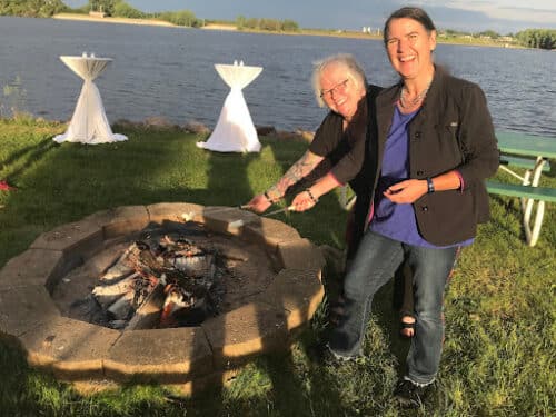  Deb Brown and Becky McCray laugh as they toast marshmallows over a fire pit by a lake at sunset. Picnic and cocktail tables are set up on the lawn.