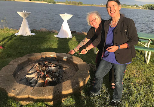 Deb Brown and Becky McCray laugh as they toast marshmallows over a fire pit by a lake at sunset. Picnic and cocktail tables are set up on the lawn.